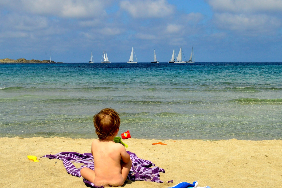 A baby sitting in the sand, looking out at the blue sea and sailboat on Menorca's north coast, near Fàvaritx.